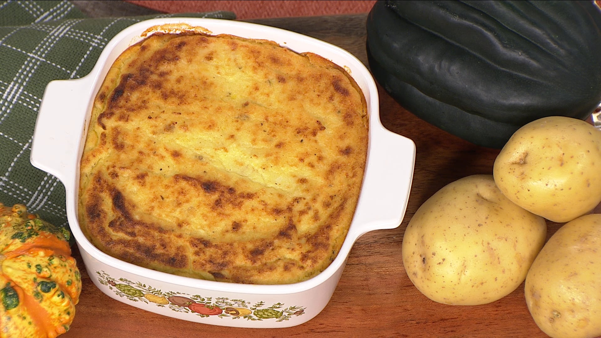 Slightly roasted and burned mash potatoes in a white casserole dish atop a wooden table with whole yellow potatoes on the side