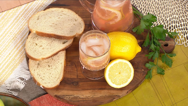 A clear glass with light pink liquid and rounded cubes of ice. A whole lemon and lemon sliced in half are beside it on the right. They are seated on a wooden circle with sliced white bread on the left-hand side.