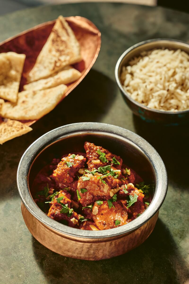 chopped chicken breast in a small brass bowl with red korma seasoning, garnished with parsley. In the upper left-hand corner, there is naan in a brass plate. On the upper right-hand corner, there is a small brass bowl of rice.