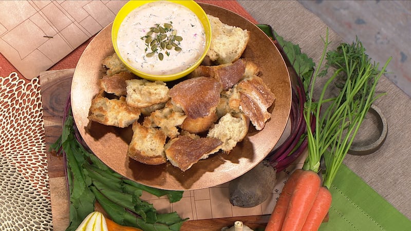 Ripped pieces of baguette lay on a circular wooden plate. In a yellow bowl at the top of this circular plate is a light pink cream dip, with pumpkin seeds as garnish.